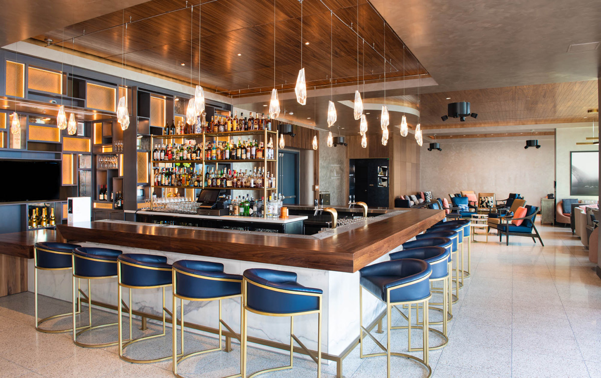 A modern bar with a long wooden counter, blue stools, and pendant lighting, with a seating area in the background.